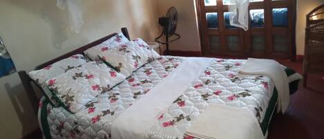 A bedroom with a bed covered in floral bedding, a wooden headboard, and a window with curtains.