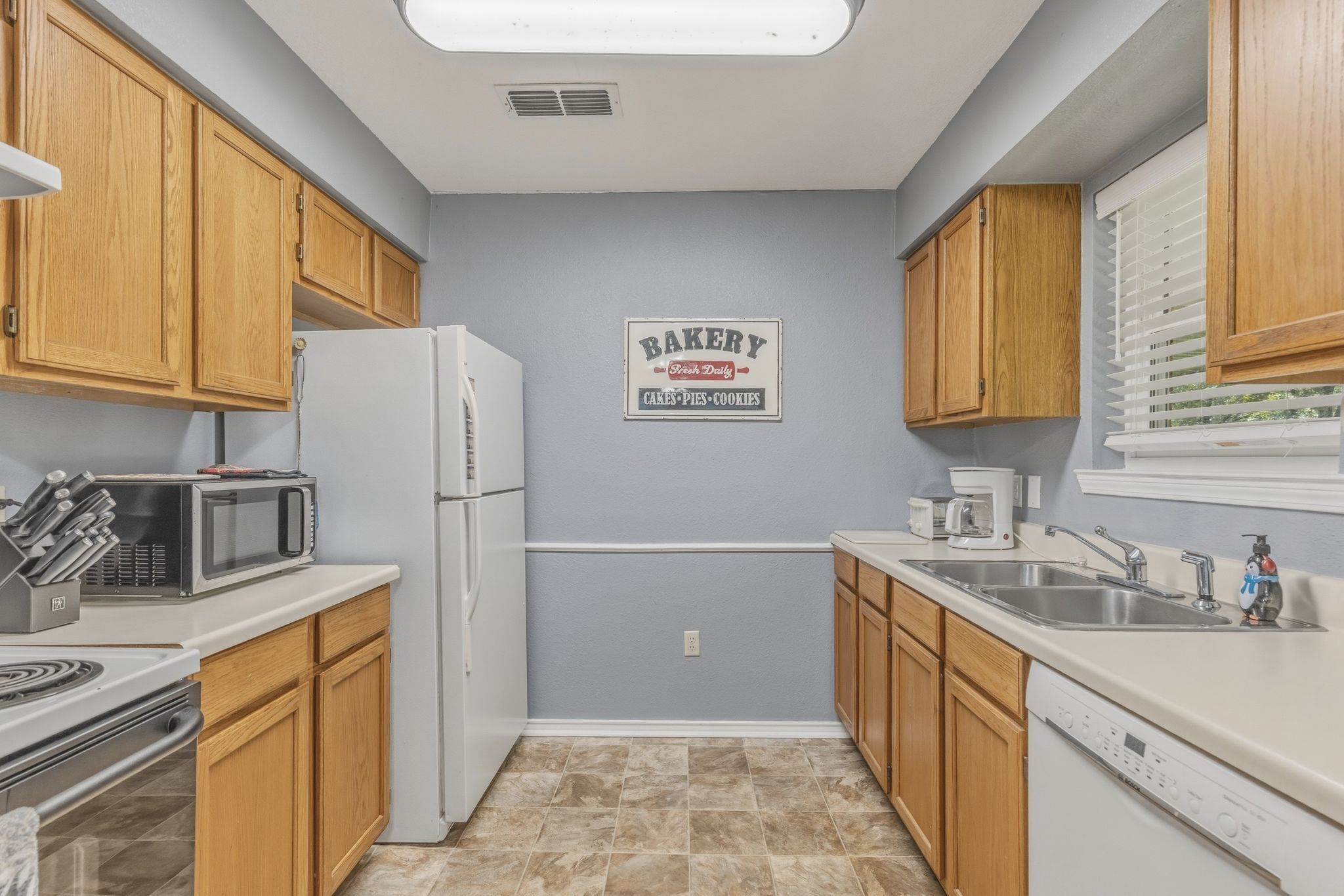 A kitchen with wooden cabinets, a white refrigerator, a microwave, a stove, a sink, and a sign that reads 'BAKERY Cakes Pies Cookies.'