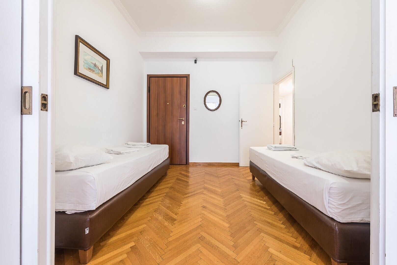 A room with two beds, a wooden herringbone floor, white walls, and a door with a circular mirror.