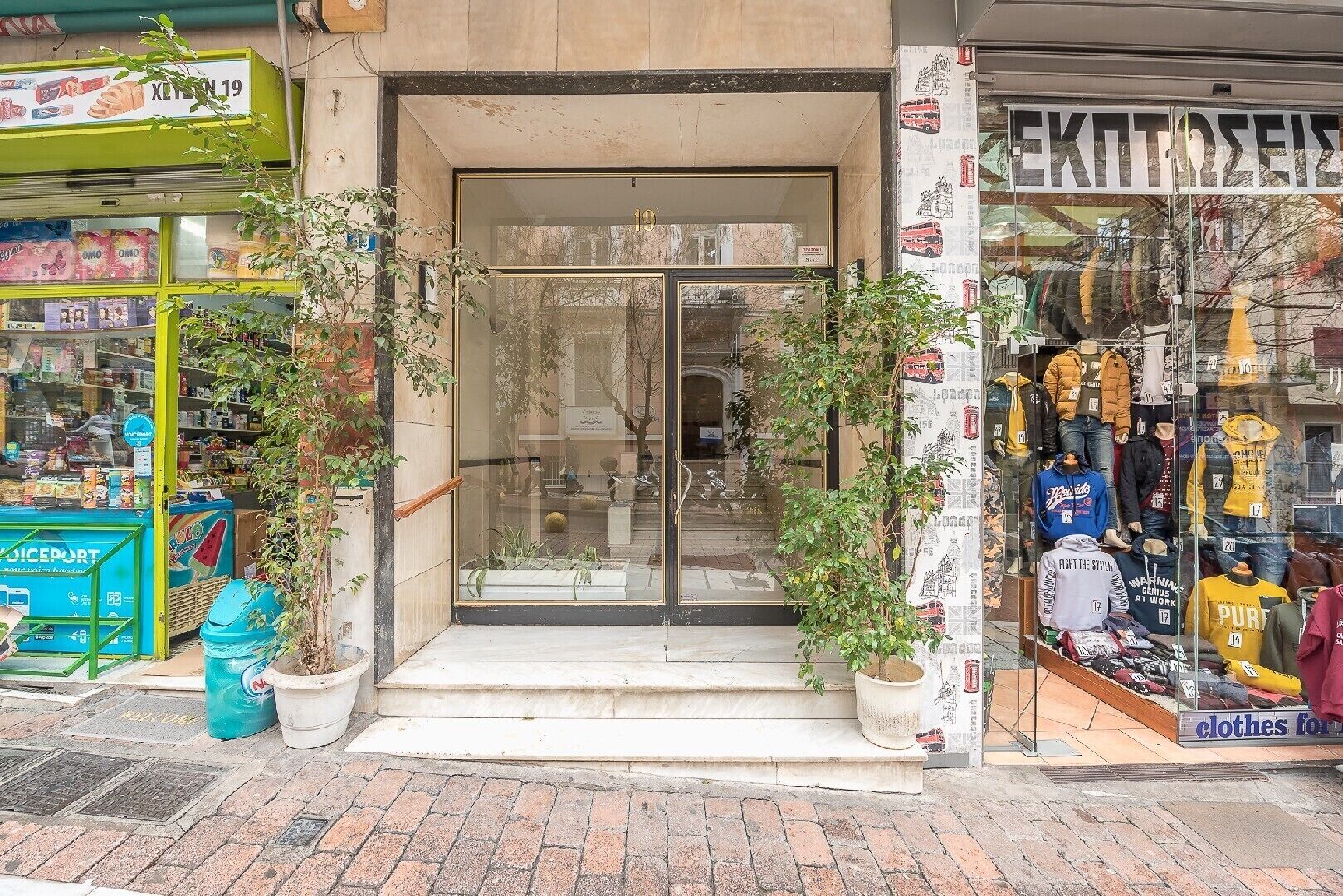 A storefront with a glass door entrance, potted plants, and various items on display including clothing and snacks.