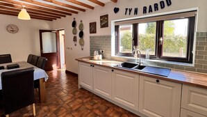 A kitchen with white cabinets, a window with a view, a clock, and a nameplate on the wall. - Ty'n Rardd Mountain Views Secure Garden Parking (Beaumaris)