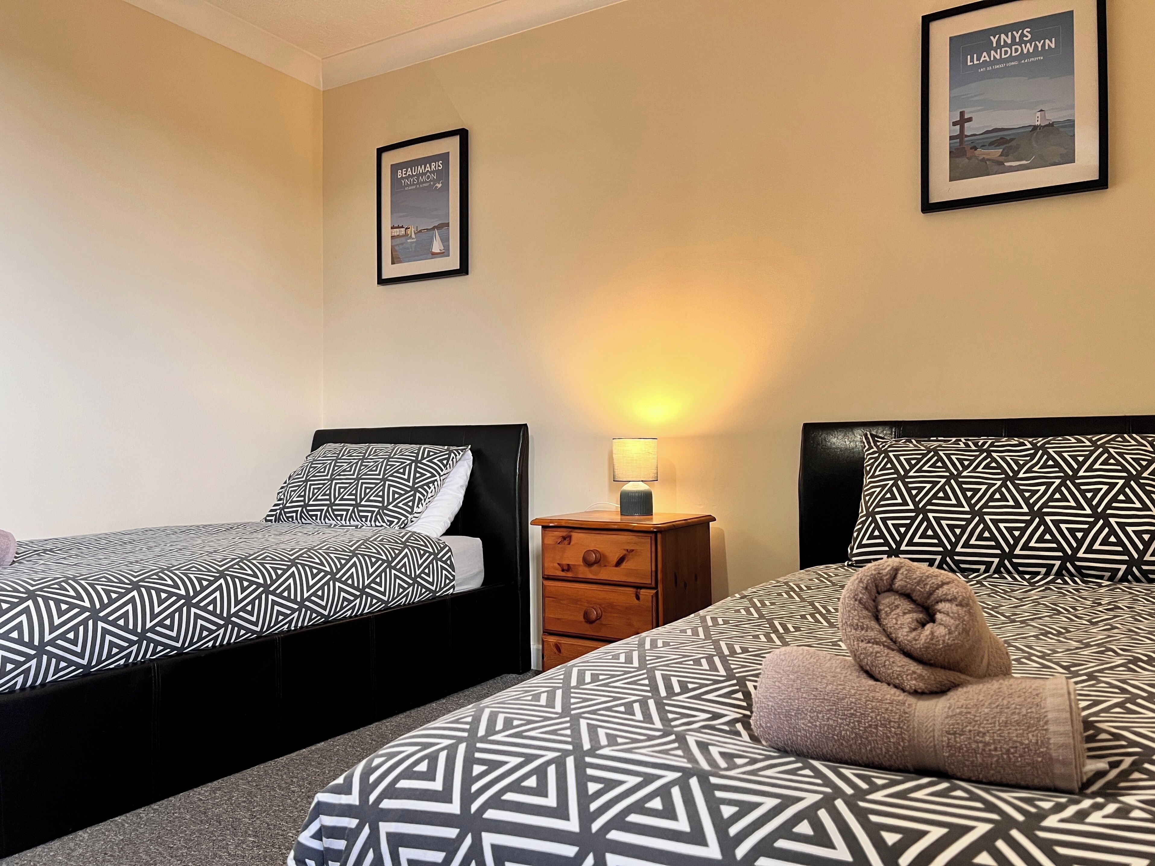 A hotel room with two beds, a nightstand with a lamp, and two framed pictures on the wall.