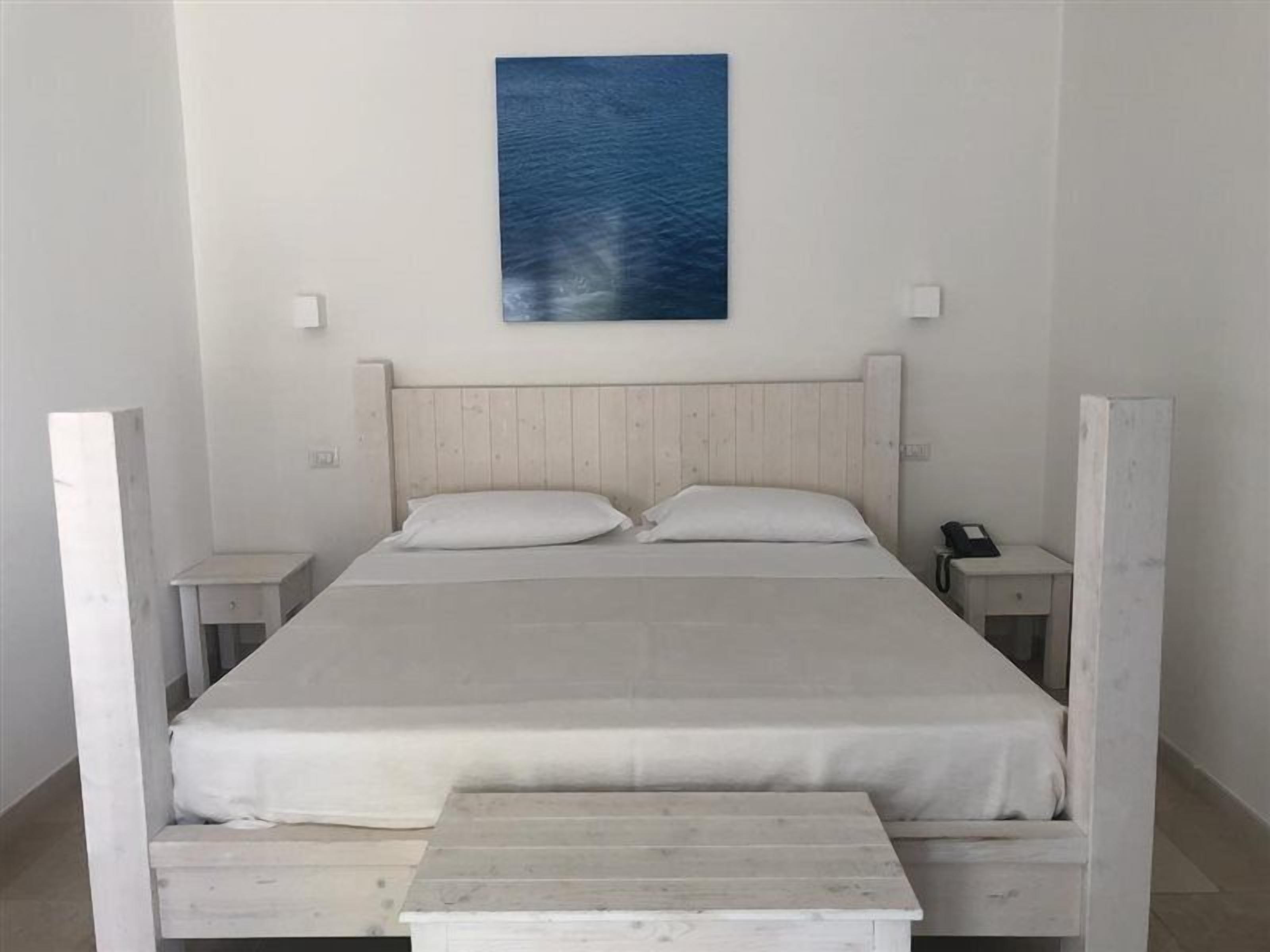 Ein schlicht eingerichtetes Schlafzimmer mit einem hölzernen Bett, weißer Bettwäsche und einem blauen Kunstwerk an der Wand.