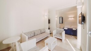 Een moderne, minimalistische woonkamer met een bank, fauteuils en een salontafel. Er is een televisie aan de muur bevestigd en een klein tafeltje met een stoel.
