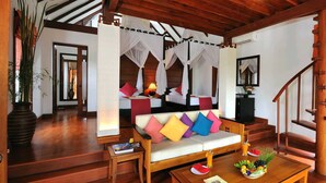 A modern hotel room with a wooden floor, a sofa with colorful cushions, a dining table with fruit, and a staircase.