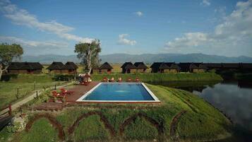 A pool with lounge chairs, a wooden deck, and thatched-roof cottages in a rural setting.