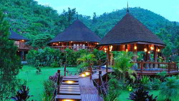A wooden pathway leading to a series of thatched-roof structures surrounded by lush greenery.
