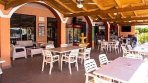 An outdoor café area with wooden tables and white chairs, a Coca-Cola refrigerator, and a ceiling fan. - Nikos Studios (Zakynthos)