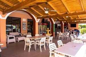 Un área de café al aire libre con mesas de madera y sillas blancas, un refrigerador de Coca-Cola y un ventilador de techo.