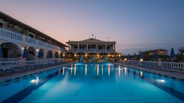 A swimming pool with lounge chairs and umbrellas, a building with a balcony, and a clear sky. - Nikos Studios (Zakynthos)