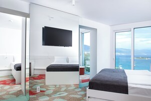 A modern hotel room with a large bed, a TV mounted on the wall, and a balcony with a view of the sea.