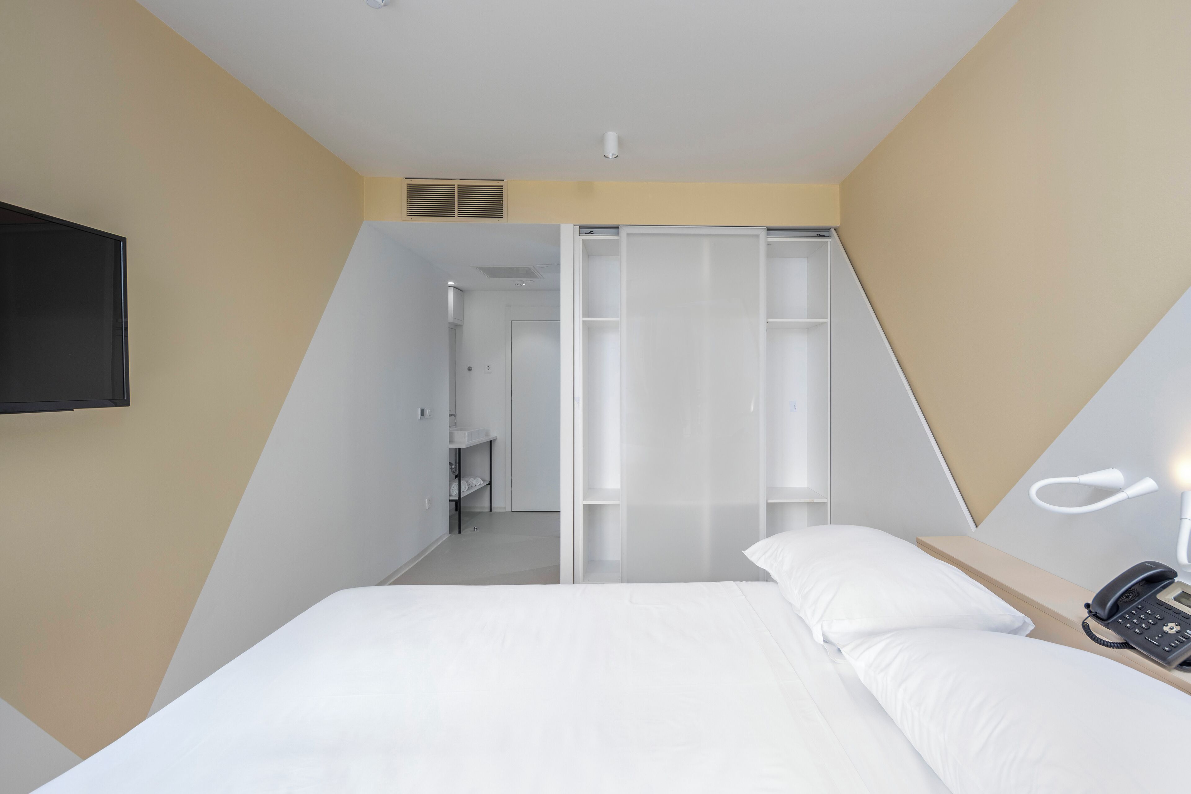 A modern hotel room with a bed, a bedside phone, and a walk-in closet with sliding doors.