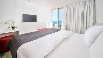 Quarto de hotel com cama, escrivaninha, cadeira, televisão e vista para o mar.