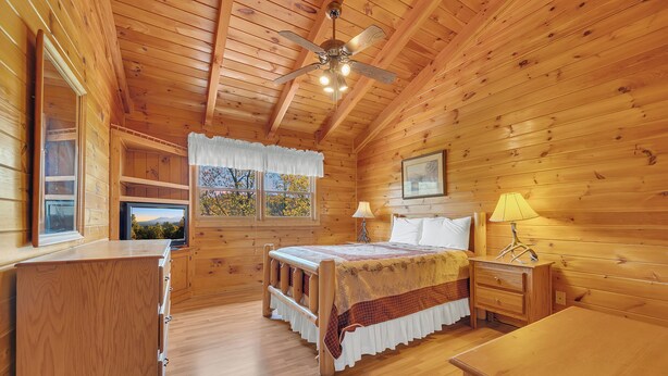 Ein Schlafzimmer in einer Holzhütte mit einem Bett, Nachttischen, einem Deckenventilator, einem Fenster mit Aussicht und einem Fernseher.