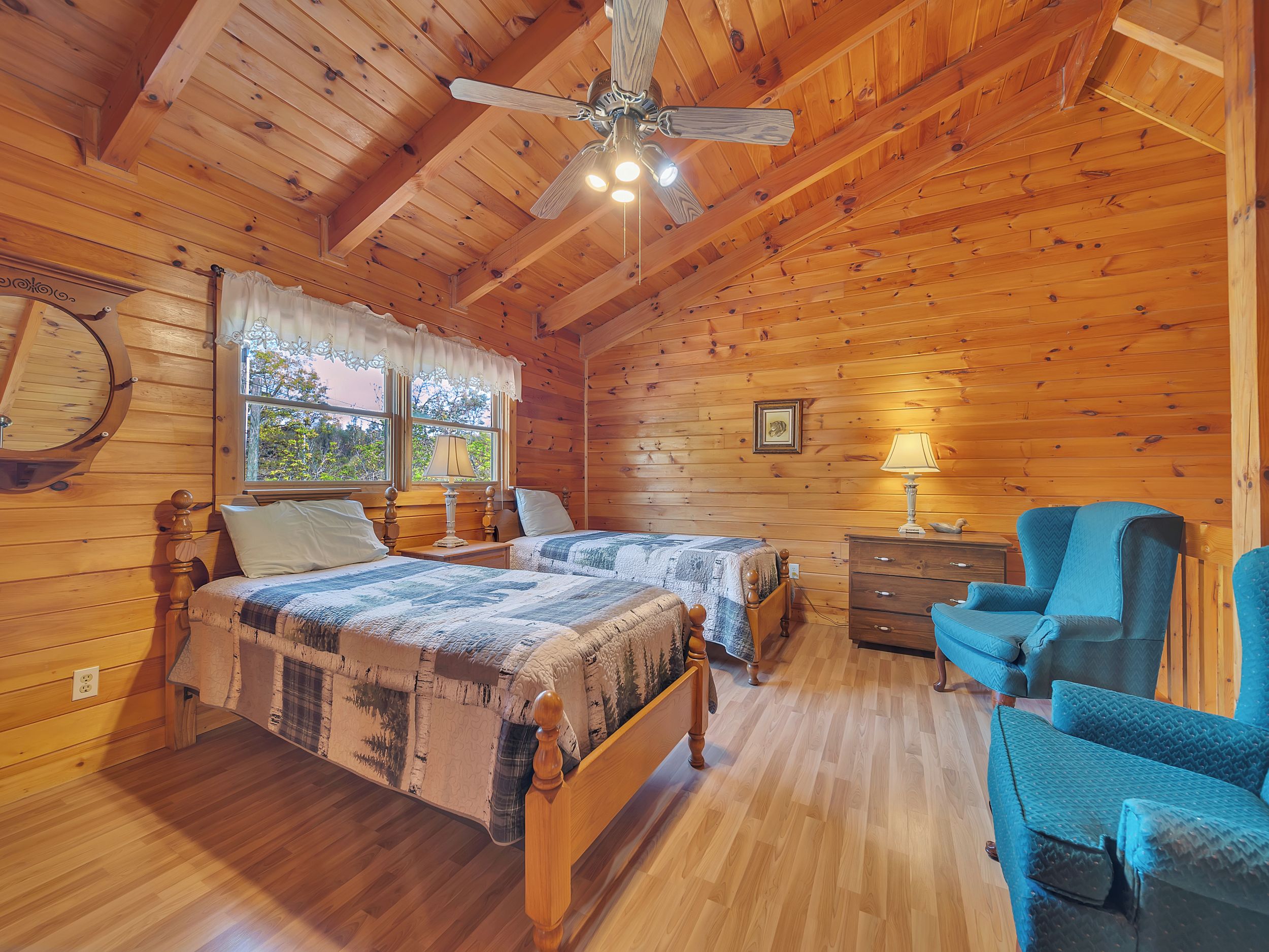 A wooden cabin bedroom with two beds, a ceiling fan, a dresser, and a blue chair.