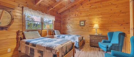 A wooden cabin bedroom with two beds, a ceiling fan, a dresser, and a blue chair.