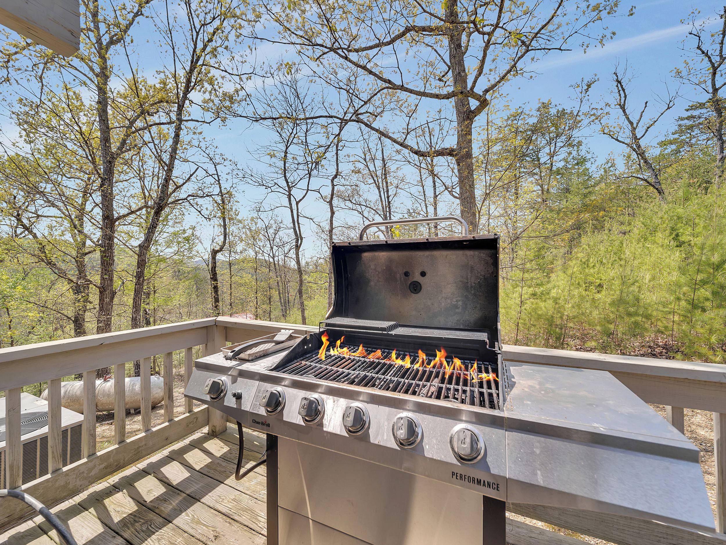 A Char-Broil gas grill with four burners, located on a wooden deck with a railing, surrounded by trees.