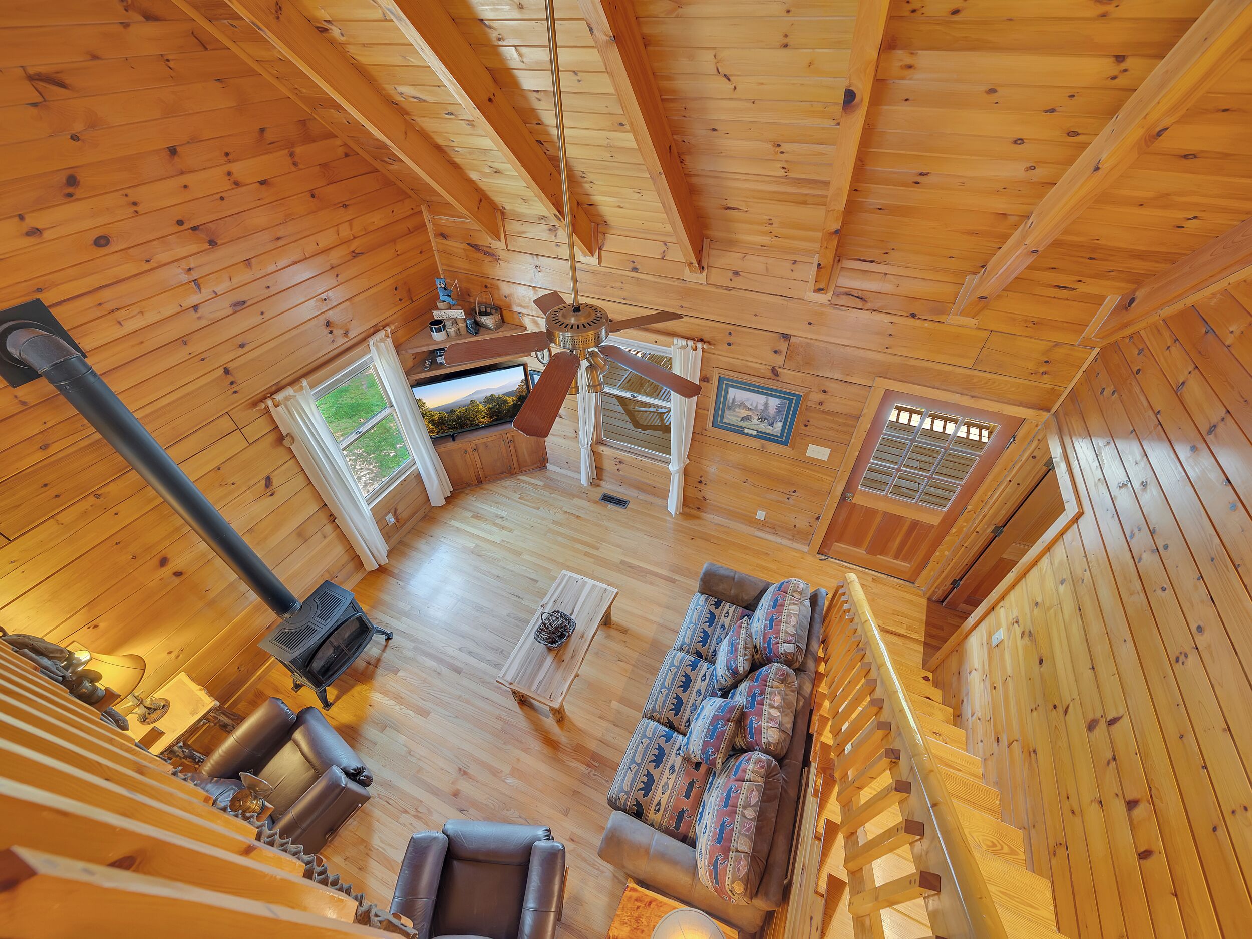A cozy cabin interior with a wooden floor, a sofa with patterned cushions, a ceiling fan, and a fireplace.