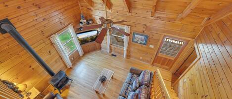 A cozy cabin interior with a wooden floor, a sofa with patterned cushions, a ceiling fan, and a fireplace.