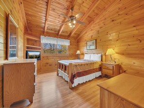 A wooden cabin bedroom with a bed, bedside tables, a ceiling fan, a window with a view, and a TV.