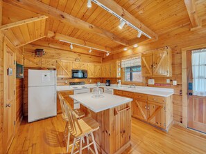 A wooden cabin-style kitchen with a refrigerator, microwave, stove, and bar stools.