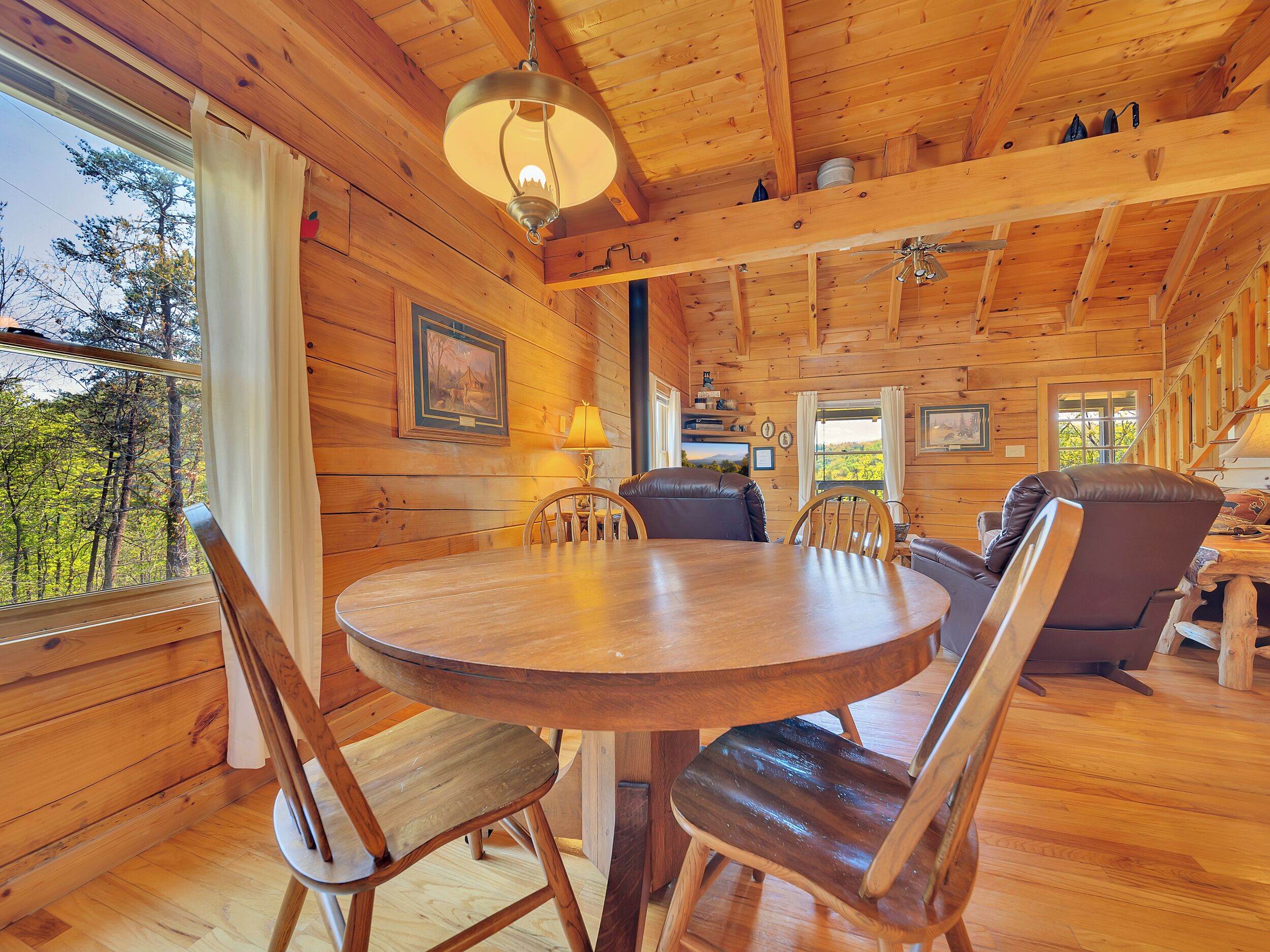 A wooden cabin interior with a round dining table, leather chairs, a hanging light fixture, and framed pictures on the walls.