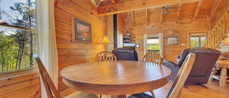 A wooden cabin interior with a round dining table, leather chairs, a hanging light fixture, and framed pictures on the walls.