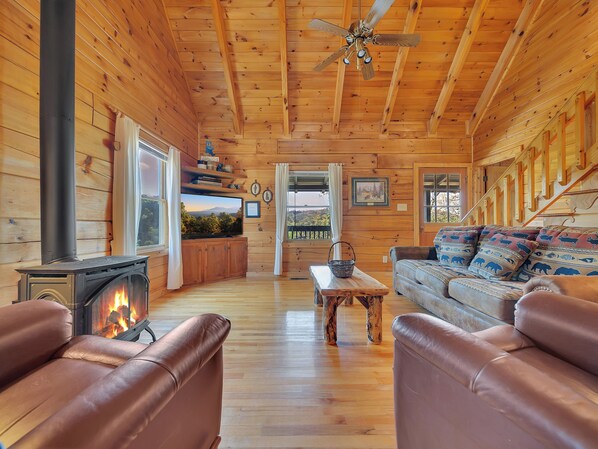 A cozy cabin interior with a fireplace, leather sofas, a wooden ceiling, and a large window overlooking a scenic landscape.