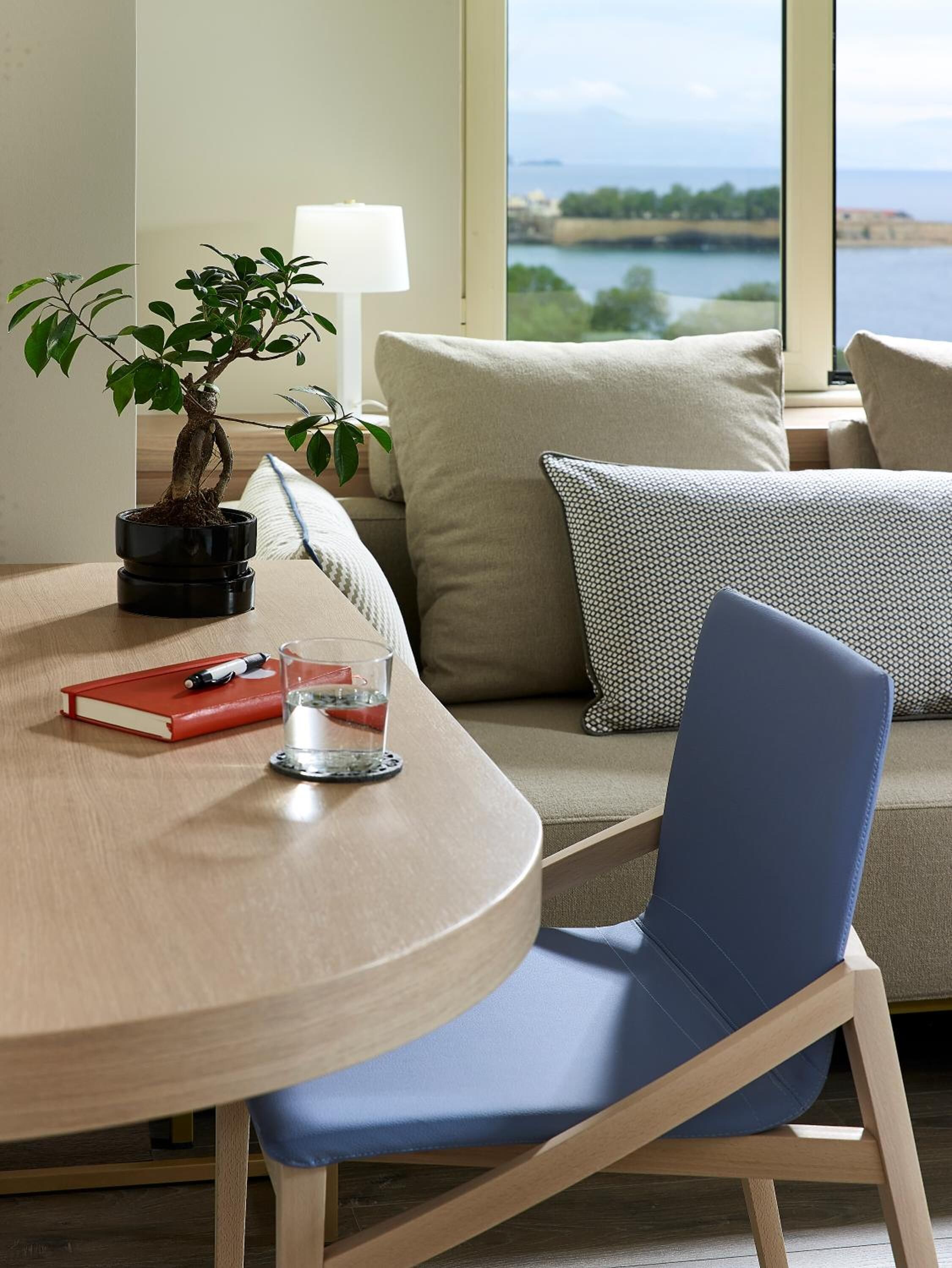 Una habitación moderna con una mesa redonda de madera, una silla azul, un sofá con cojines estampados, una planta en maceta y vistas a una masa de agua a través de grandes ventanales.