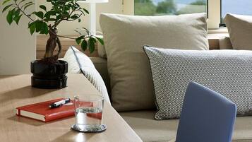 Una habitación moderna con una mesa redonda de madera, una silla azul, un sofá con cojines estampados, una planta en maceta y vistas a una masa de agua a través de grandes ventanales.