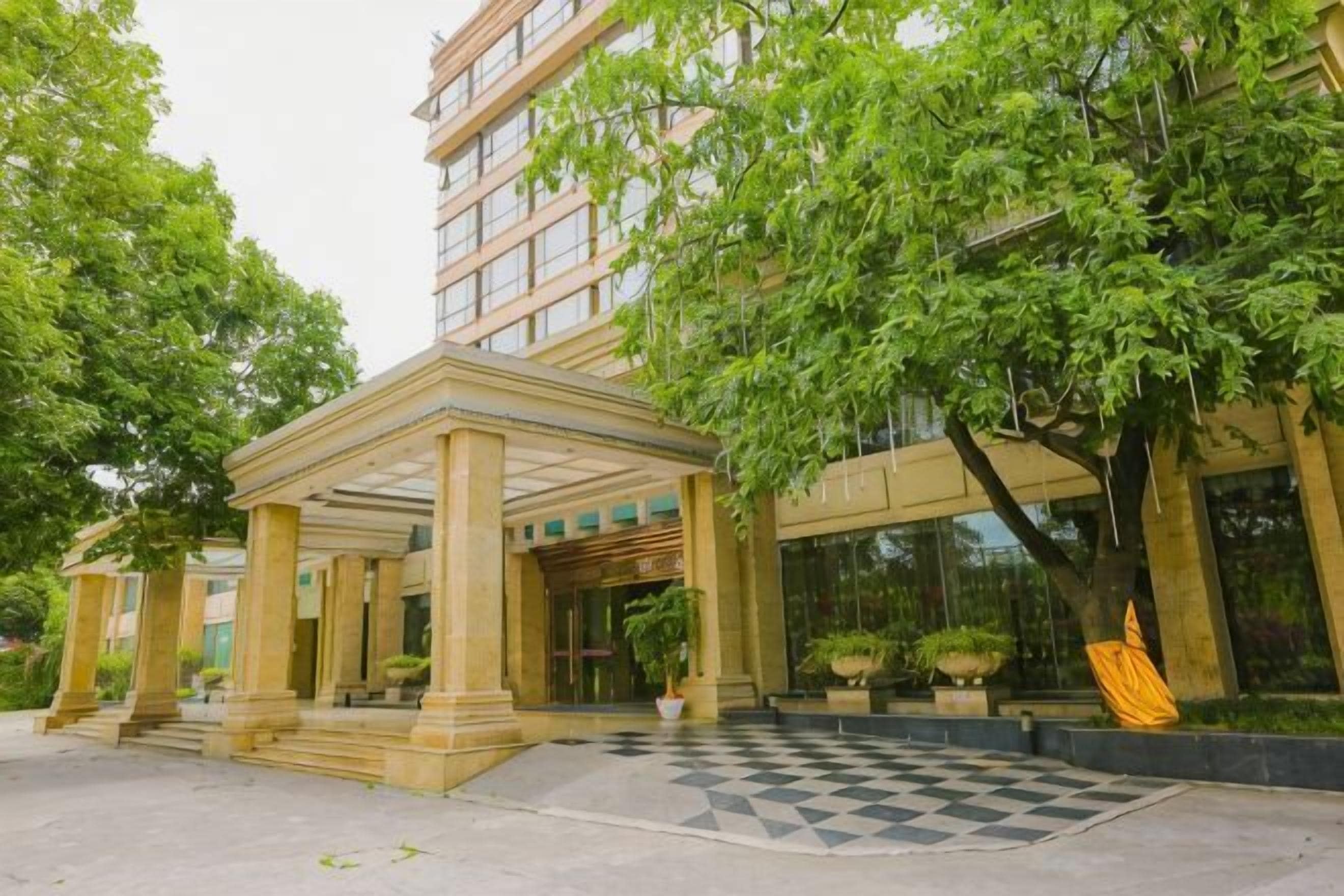 A hotel entrance with a covered patio, a checkered pavement, and lush greenery.