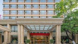 Entrada de un hotel con toldo rojo, grandes columnas y una cubierta de vidrio.