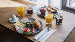 A breakfast table with croissants, sliced bread, cheese, tomatoes, cucumbers, a croissant, a plate with a croissant, a glass of orange juice, a glass of coffee, and a glass of juice. - Railway Apartments (Vilnius)