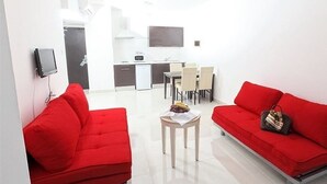 A modern living room with red sofas, a small table with fruits, and a kitchen area in the background. - Meandros Hotel Apts (Protaras)