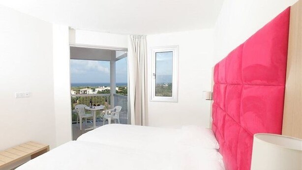 A hotel room with a large bed, a balcony with a table and chairs, and a view of the city. - Meandros Hotel Apts (Protaras)