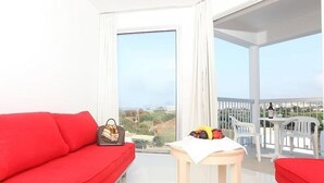 A bright room with a red sofa, a small table with fruits, and a view of greenery and buildings through large windows. - Meandros Hotel Apts (Protaras)