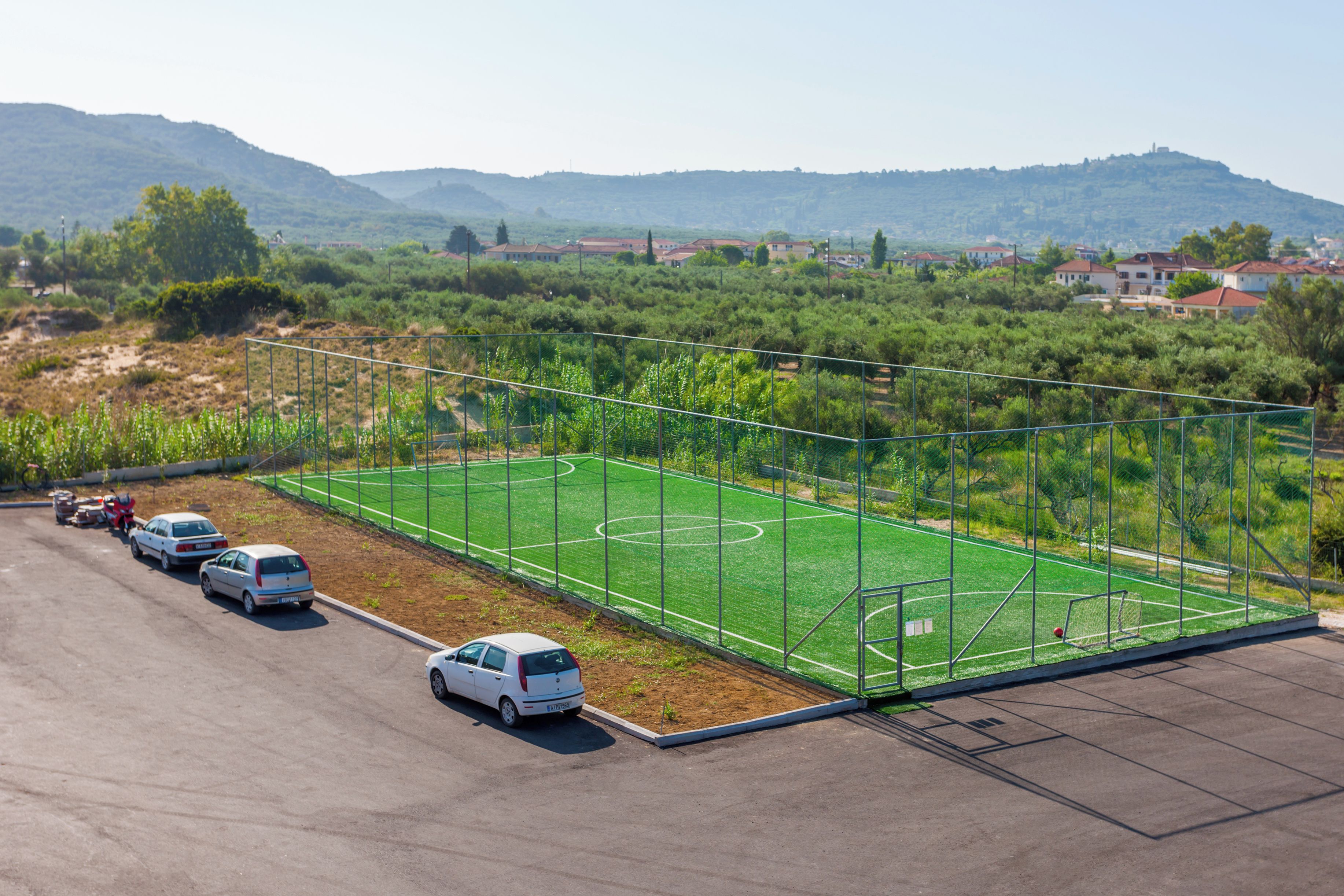 Ein eingezäuntes Sportfeld mit einem Parkplatz und einem Bergpanorama im Hintergrund.