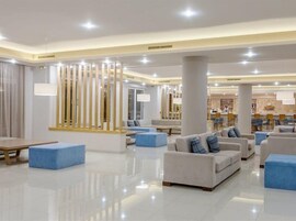 Un vestíbulo amplio con diseño moderno, que cuenta con una zona de estar central con sofás y otomanas azules, un mostrador de recepción y un área de comedor al fondo.