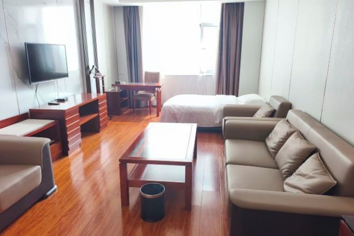 A modern hotel room with a wooden floor, a bed, a sofa, a TV, a desk, and a window with curtains.