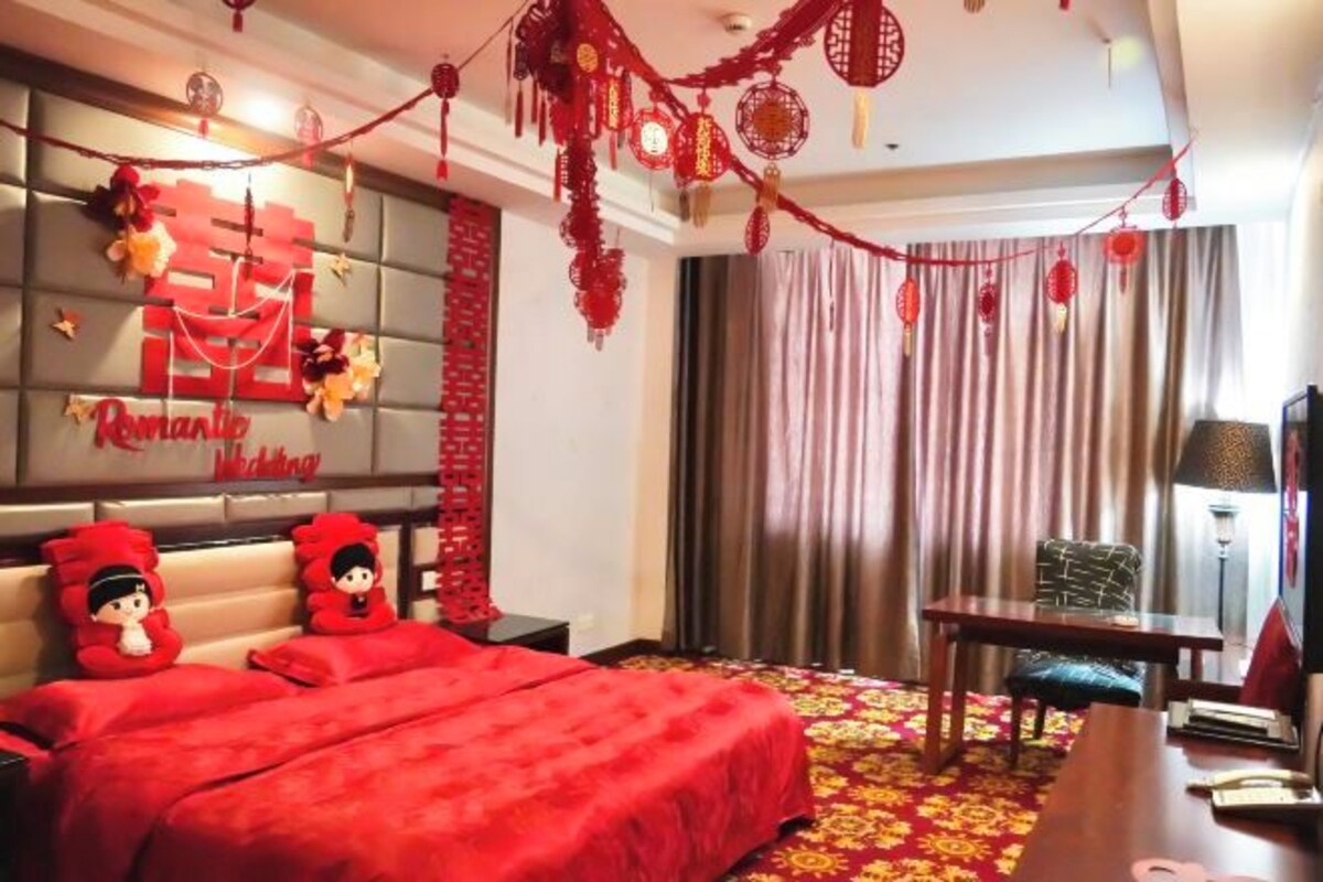 A hotel room with a red bedspread, a wall with a 'Romantic Wedding' sign, a table, and a chair.