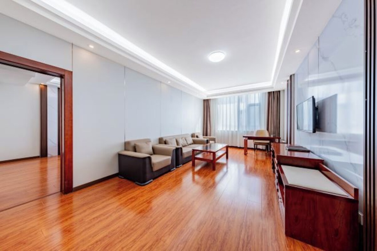 A modern hotel room with a wooden floor, a sofa set, a dining table, and a TV mounted on the wall.
