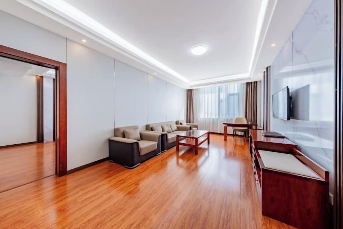 Una habitación de hotel moderna con piso de madera, un conjunto de sofás, una mesa de comedor y un televisor montado en la pared.