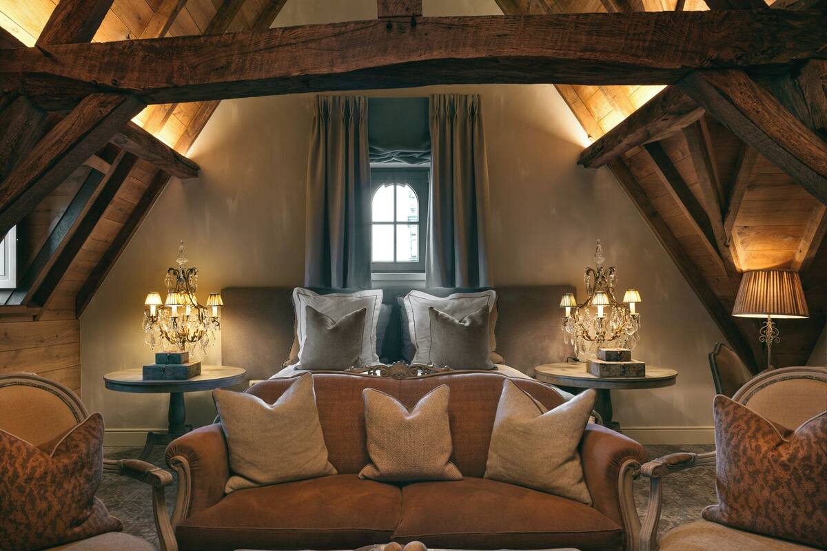 A cozy attic room with a brown sofa, two side tables with lamps, and a large window with curtains.