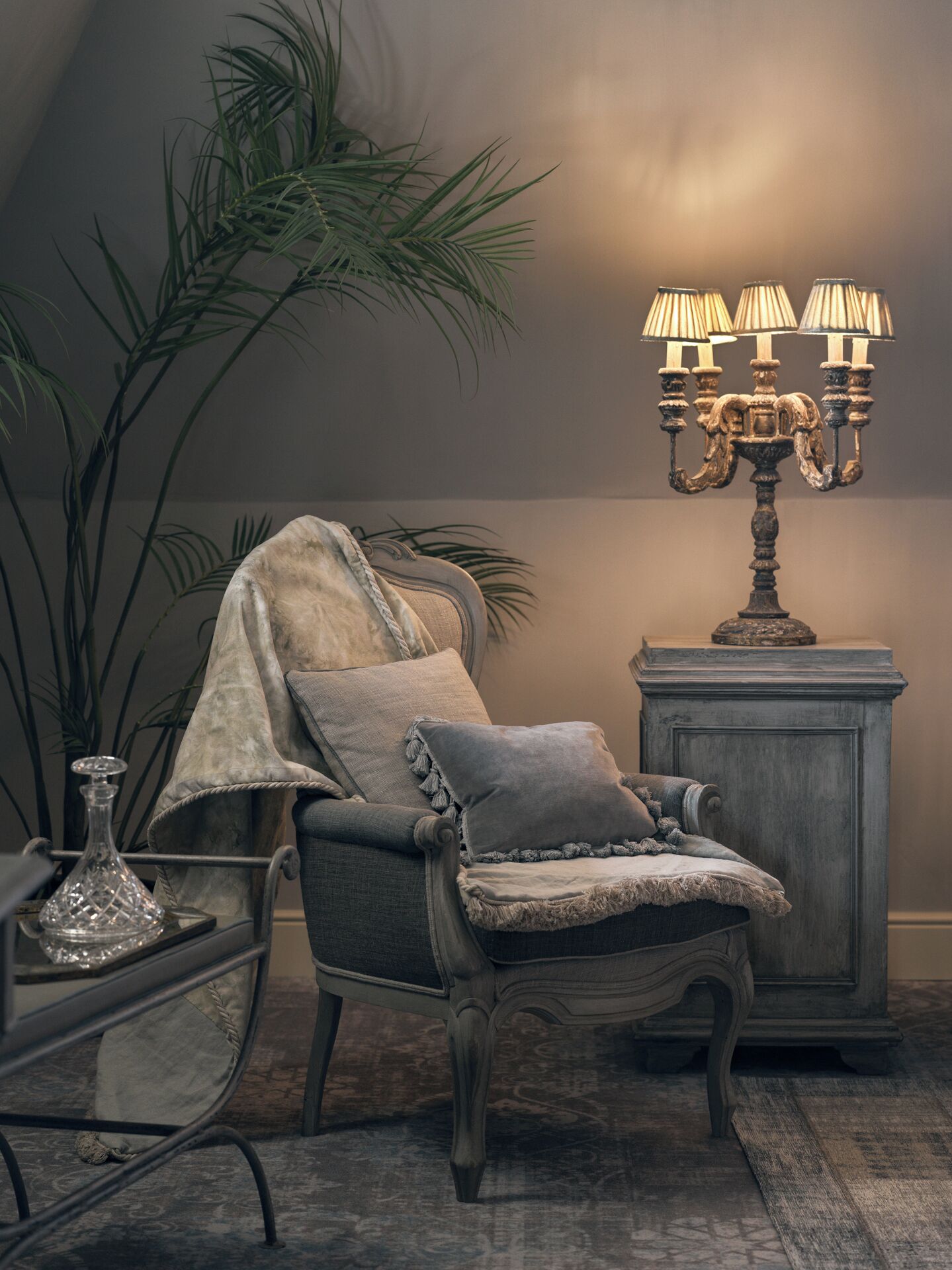 A chair with a blanket, a lamp, and a plant in a room with a patterned carpet.