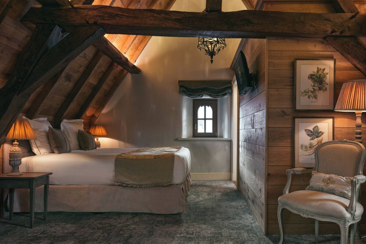 A cozy attic bedroom with a bed, a chair, a window, and framed artwork on the walls.