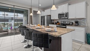 A modern kitchen with white cabinets, a granite countertop, and stainless steel appliances. There are bar stools and a dining area with a view of the outside. - 5BD Windsor Westside Private Pool (WW 8920) (Kissimmee)
