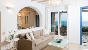 A modern living room with a beige sofa, a glass coffee table, and a view of the sea through a large sliding glass door. - Exquisite "Villa Luna" with Pool & Jacuzzi close to the amazing Lia beach! (Mykonos)