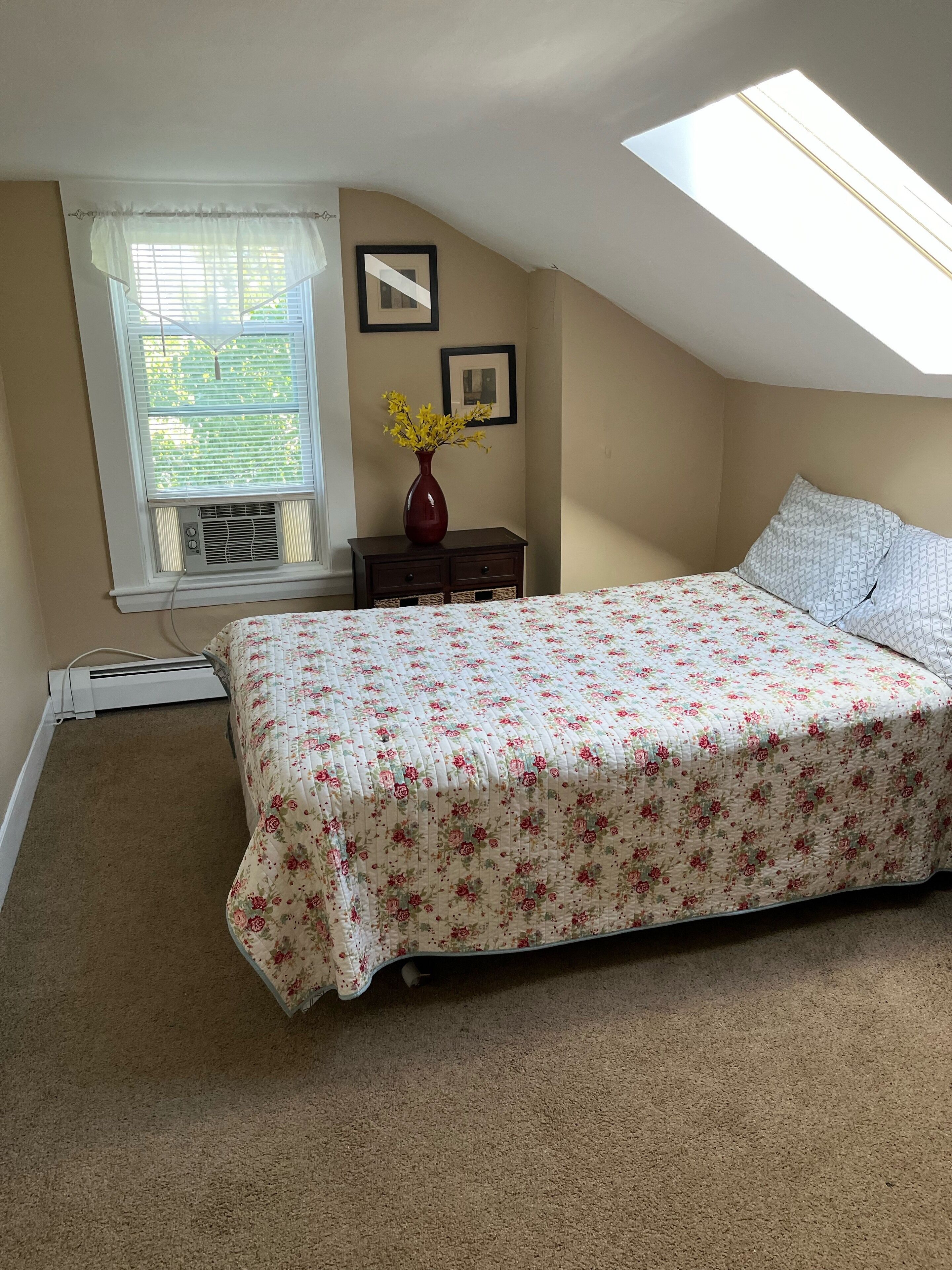A bedroom with a sloped ceiling, a skylight, a bed with floral bedding, a nightstand with a vase, and a window with blinds.