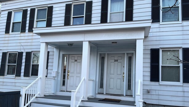 A two-story white house with black shutters, a front porch, and a brick driveway. - Spacious Apt. 2 car free parking great location. (Cambridge)