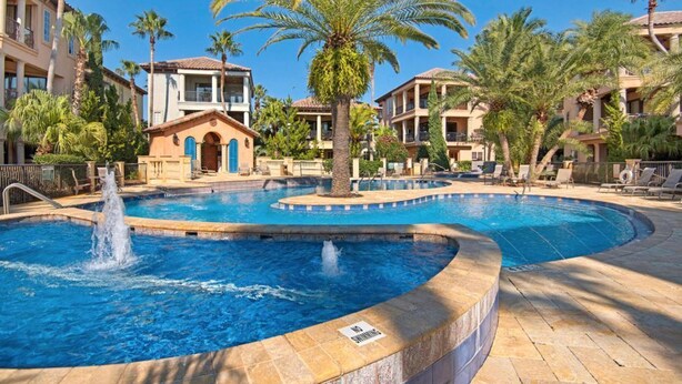 A pool area with a fountain, surrounded by palm trees and buildings with balconies.